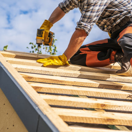 Do-It-Yourself RentalsA construction worker in a plaid shirt and orange pants uses a power drill on a wooden roof under a blue sky. The scene conveys focus and craftsmanship.