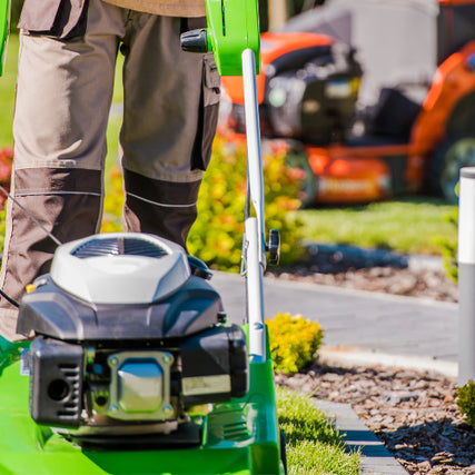 Landscaping RentalsA person in brown pants operates a green lawnmower on a sunny day. Vibrant green grass and colorful shrubs are visible, with another mower blurred in the background.