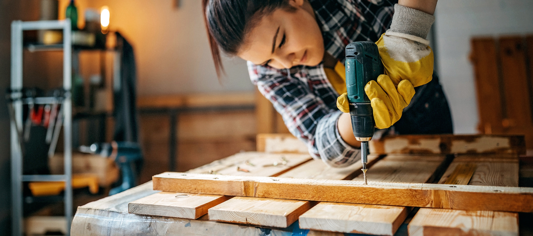 A woman in a plaid shirt and yellow gloves uses a drill on a wooden plank in a workshop. The scene conveys focus, craftsmanship, and determination.