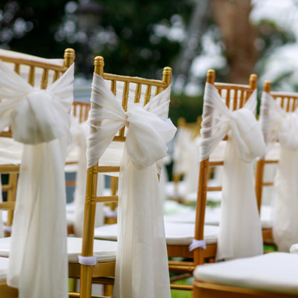 Party & Event RentalsRows of elegant gold chairs adorned with white chiffon bows set up outdoors, conveying a serene and romantic wedding atmosphere.